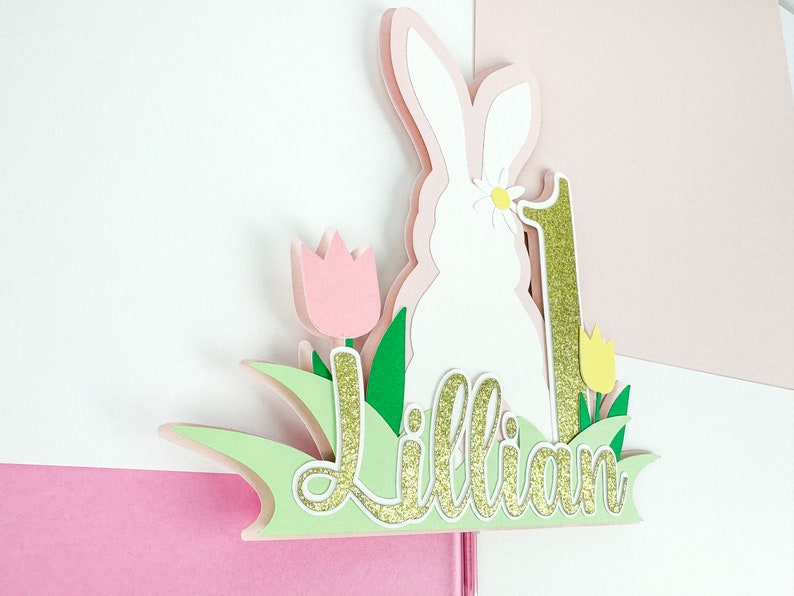 May include: A white bunny cake topper with a pink flower, green leaves, and a gold glitter number one. The name "Lillian" is written in gold glitter on a green background.