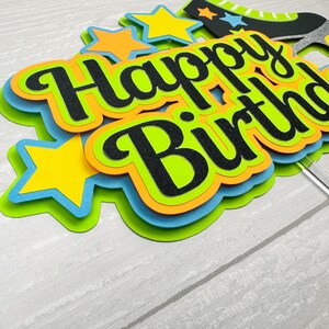 May include: A layered paper cake topper featuring the black glitter text Happy Birthday. The design includes bright yellow, orange, light blue, and lime green star shapes and a black roller skate accent, all attached to a clear plastic stick.