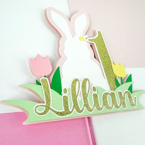 May include: A green, pink, and gold paper cake topper with a white bunny, a number one, and the name "Lillian".