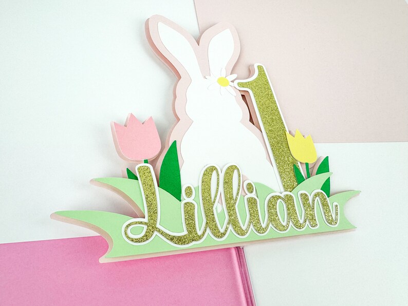May include: A green, pink, and gold paper cake topper with a white bunny, a number one, and the name "Lillian".