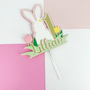 May include: A white bunny cake topper with pink ears and a gold glitter number one. The bunny is surrounded by green grass and yellow flowers. The name "Lillian" is written in gold glitter on a green banner.
