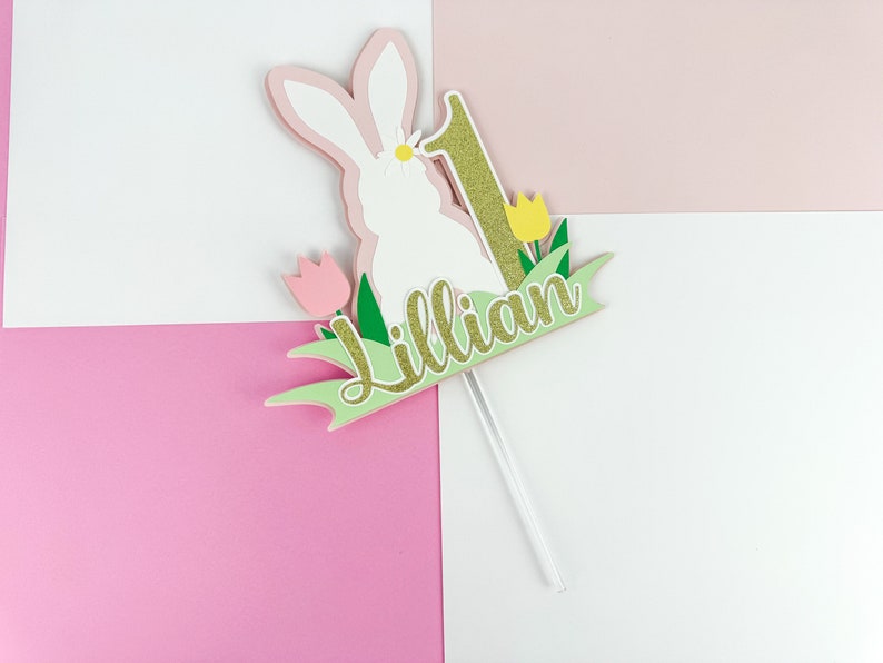 May include: A white bunny cake topper with pink ears and a gold glitter number one. The bunny is surrounded by green grass and yellow flowers. The name "Lillian" is written in gold glitter on a green banner.