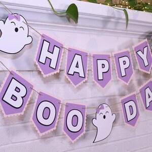 May include: A festive Halloween banner with the words "HAPPY BOO DAY" in white block letters on lavender pennants with pink scalloped edges. The banner is decorated with smiling ghost cutouts and strung on twine.