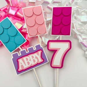 May include: A set of five cake toppers featuring a purple and pink castle with the name "Abby" on it, a pink number seven, and three pink and blue Lego-style blocks.