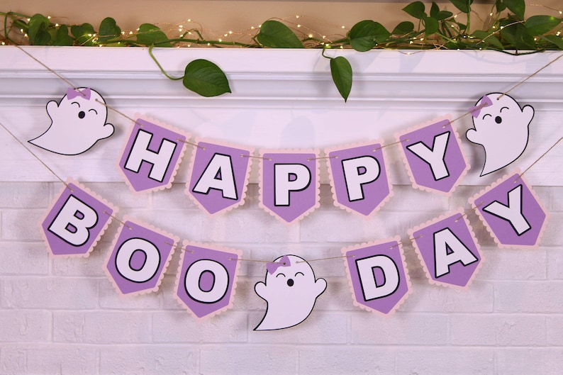 May include: A Halloween banner with the words "HAPPY BOO DAY" in white letters on purple pennants. The pennants are trimmed with a scalloped edge and strung with twine. White ghost cutouts are also attached.
