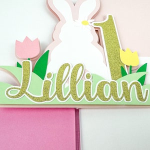 May include: A white bunny cake topper with a pink flower on its head, surrounded by green grass, pink and yellow tulips, and the name "Lillian" in gold glitter. The number "1" is also in gold glitter.