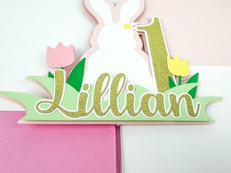 May include: A white bunny cake topper with a pink flower on its head, surrounded by green grass, pink and yellow tulips, and the name "Lillian" in gold glitter. The number "1" is also in gold glitter.