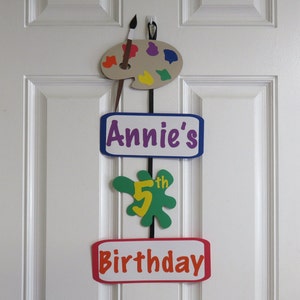 May include: A colorful birthday decoration for a child's 5th birthday. The decoration features a paint palette, a paint splatter with the number 5, and the words "Annie's 5th Birthday".