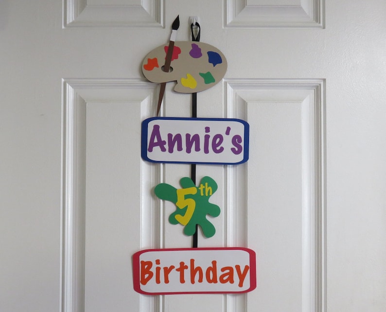 May include: A colorful birthday decoration for a child's 5th birthday. The decoration features a paint palette, a paint splatter with the number 5, and the words "Annie's 5th Birthday".