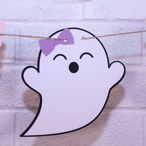 May include: A white ghost-shaped banner with a purple bow and a smiling face. The banner is strung on twine and has purple and pink scalloped edges. The background is a white brick wall.