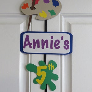 May include: A colorful paint palette, paintbrush, and paper cutouts spelling "Annie's 5th Birthday" hanging on a white door.