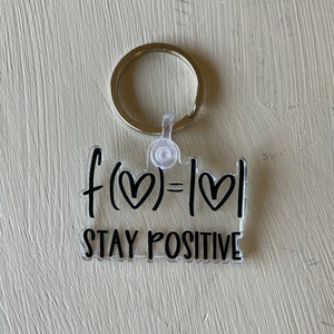 May include: Clear acrylic keychain with a silver keyring. The keychain features black text that reads "f(heart) = |heart|" and "STAY POSITIVE". The design includes heart symbols.