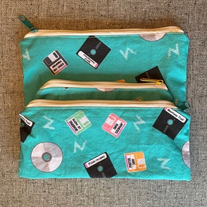 May include: Three turquoise zippered pouches with a repeating pattern of floppy disks and CDs. The pouches have white zippers and gold zipper pulls. The floppy disks have various labels, including "Science Fair", "Grandma's Cat Photos", "Japanese ROMs", "Photos 1989", and "Pizza Recipes".