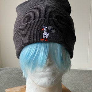 Puede incluir: Un gorro gris oscuro con un pequeño diseño bordado de un ganso blanco que lleva un objeto azul y blanco. El ganso tiene patas naranjas.