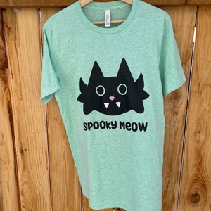 May include: A light green t-shirt featuring a black cartoon cat with fangs and the text "spooky meow" printed on the front.