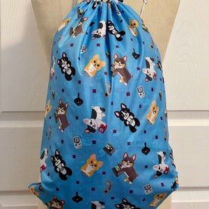 May include: A blue drawstring bag with a pattern of cartoon corgis playing video games. The corgis are in various colors and poses, and the video game controllers are in different styles. The bag has black drawstrings.
