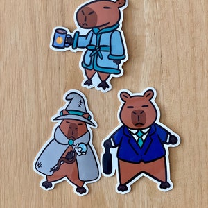 May include: Three capybara stickers with different outfits. One is wearing a blue robe and holding a mug, another is wearing a gray wizard robe and holding a wand, and the last one is wearing a blue suit and holding a briefcase.