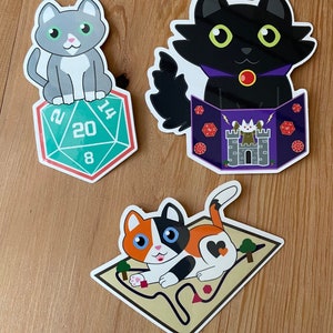 May include: Three cat stickers with different designs. The first sticker features a gray and white cat sitting on top of a green 20-sided die. The second sticker features a black cat wearing a purple collar and holding a book with a castle and dice on it. The third sticker features a calico cat on a brown map with a red heart.
