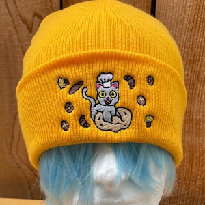 May include: Mustard yellow beanie with a rolled cuff. The beanie features an embroidered design of a gray cat wearing a chef's hat, surrounded by baked goods. The cat is sitting on a donut. The beanie is displayed on a mannequin head with blue hair.