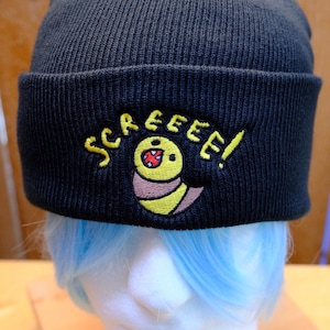 Peut inclure: Bonnet en tricot noir avec le mot "SCREEEE!" brodé en jaune, accompagné d'un dessin de ver de dessin animé. Le bonnet est présenté sur une tête de mannequin avec des cheveux bleus.