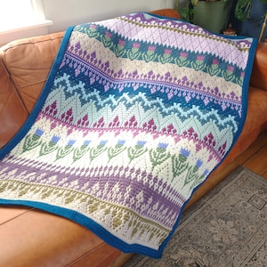 May include: A rectangular crochet blanket featuring a repeating pattern of thistle motifs and geometric bands in shades of teal, lavender, cream, and deep purple. The blanket is finished with a solid dark teal border along all four edges.