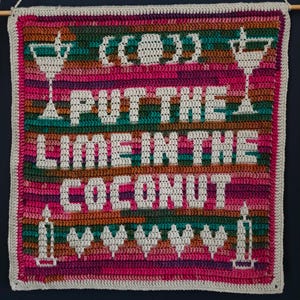 Puede incluir: Un tapiz de ganchillo cuadrado con un fondo de hilo jaspeado en rosa, verde y marrón. La pieza muestra el texto blanco "PUT THE LIME IN THE COCONUT" con iconos gráficos blancos de dos copas de cóctel, un coco y cinco corazones.