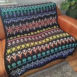 K&ouml;nnte beinhalten: Eine schwarze H&auml;keldecke mit einem farbenfrohen geometrischen Muster. Das Muster zeigt verschiedene Formen, darunter Dreiecke, Quadrate und Rauten. Die Decke ist aus weichem, strukturiertem Garn gefertigt und hat einen Fransenrand.