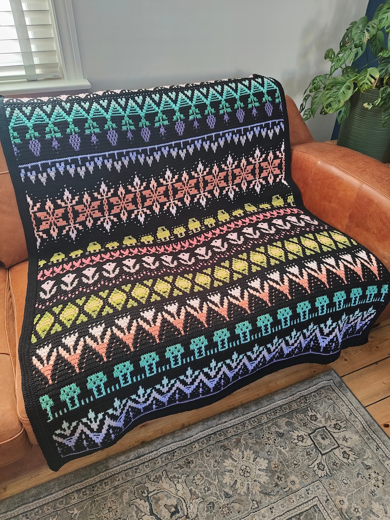 K&ouml;nnte beinhalten: Eine schwarze H&auml;keldecke mit einem farbenfrohen geometrischen Muster. Das Muster zeigt verschiedene Formen, darunter Dreiecke, Quadrate und Rauten. Die Decke ist aus weichem, strukturiertem Garn gefertigt und hat einen Fransenrand.