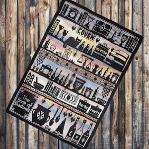 May include: Black and white crochet blanket with a colourful ombre design. The blanket features a variety of witchy symbols and text, including "The Book of Shadows", "Coven", "Herbs", "Roots", and "Magic".