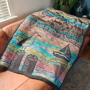 May include: A colorful, crocheted blanket featuring a coastal town scene. The blanket showcases a vibrant palette of blues, pinks, and yellows, with a dark gray border. The design includes a town, a boat, and other architectural elements.