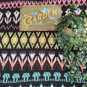 K&ouml;nnte beinhalten: Eine schwarz-wei&szlig;e H&auml;keldecke mit einem bunten geometrischen Muster. Ein Metallschild mit dem Text "Abi's Garden Sit-Relax Enjoy Where love is grown & memories are made" liegt auf der Decke. Rechts im Bild befindet sich eine Topfpflanze.