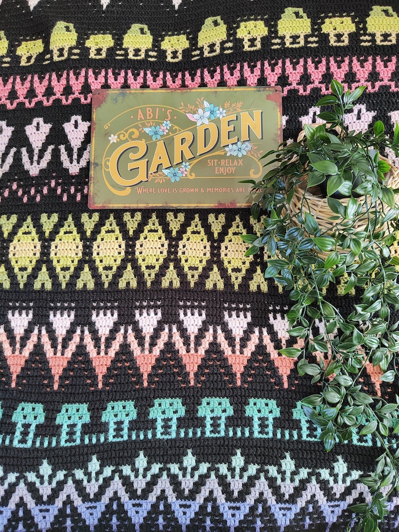 K&ouml;nnte beinhalten: Eine schwarz-wei&szlig;e H&auml;keldecke mit einem bunten geometrischen Muster. Ein Metallschild mit dem Text "Abi's Garden Sit-Relax Enjoy Where love is grown & memories are made" liegt auf der Decke. Rechts im Bild befindet sich eine Topfpflanze.
