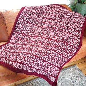 Puede incluir: Una manta de ganchillo burdeos y blanca con un patrón geométrico. La manta está sobre un sofá de cuero marrón. El patrón es un diseño repetitivo de líneas blancas sobre un fondo burdeos. La manta tiene un borde burdeos liso.