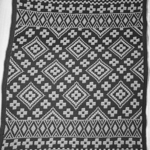 May include: A black and gray crocheted blanket with a geometric pattern. The blanket has a diamond pattern with smaller flower-like shapes in the center of each diamond.
