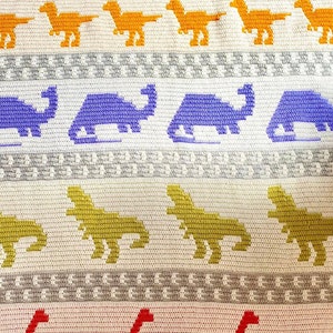 May include: A white crocheted blanket with a pattern of colorful dinosaurs. The dinosaurs are in rows and include pink, orange, purple, yellow, and green colors.