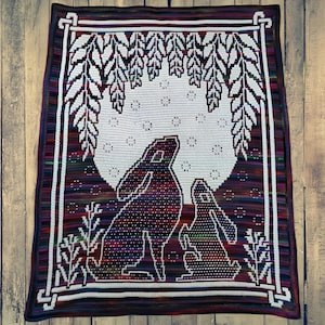 May include: A crocheted blanket featuring a design of two rabbits silhouetted against a full moon. The blanket has a white border with a repeating pattern and a multicolored striped edge. The rabbits are in a field of grass.