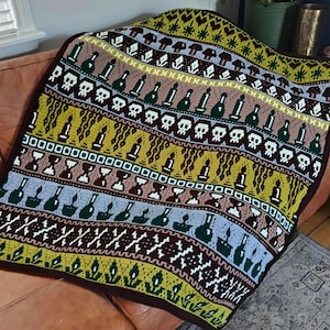May include: A colorful, crocheted blanket with a repeating pattern of skulls, bottles, candles, and hourglasses. The blanket features a dark brown border and a variety of colors including green, yellow, brown, and blue. The blanket is draped over a brown couch.