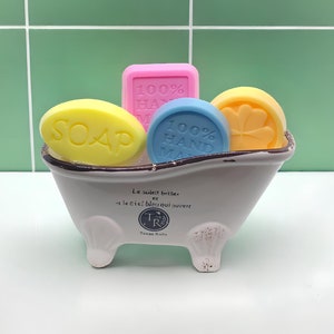 May include: A white ceramic bathtub with four feet, filled with four colorful bars of soap. The soap bars are yellow, pink, blue, and orange. The bathtub has the text "Le soleil briller et le ciel bleu qui ouvert" and "Temps Riche" printed on the side.