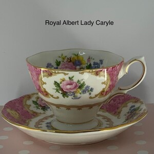 May include: A Royal Albert Lady Caryle teacup and saucer set featuring a white porcelain base with pink floral panels, gold trim, and a colourful bouquet of roses and blue flowers. The text Royal Albert Lady Caryle is printed above the cup.