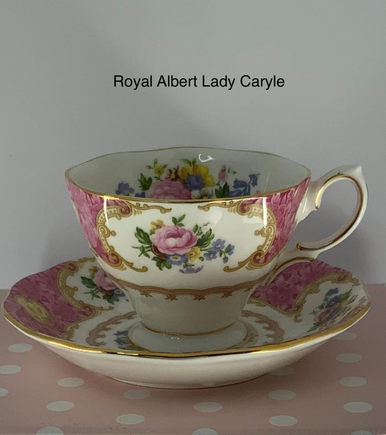 May include: A Royal Albert Lady Caryle teacup and saucer set featuring a white porcelain base with pink floral panels, gold trim, and a colourful bouquet of roses and blue flowers. The text Royal Albert Lady Caryle is printed above the cup.