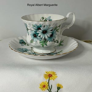 May include: A Royal Albert Marguerite teacup and saucer set featuring a white porcelain base with a blue daisy floral pattern and gold trim. The set is displayed above a white fabric cloth embroidered with three yellow daisy flowers.