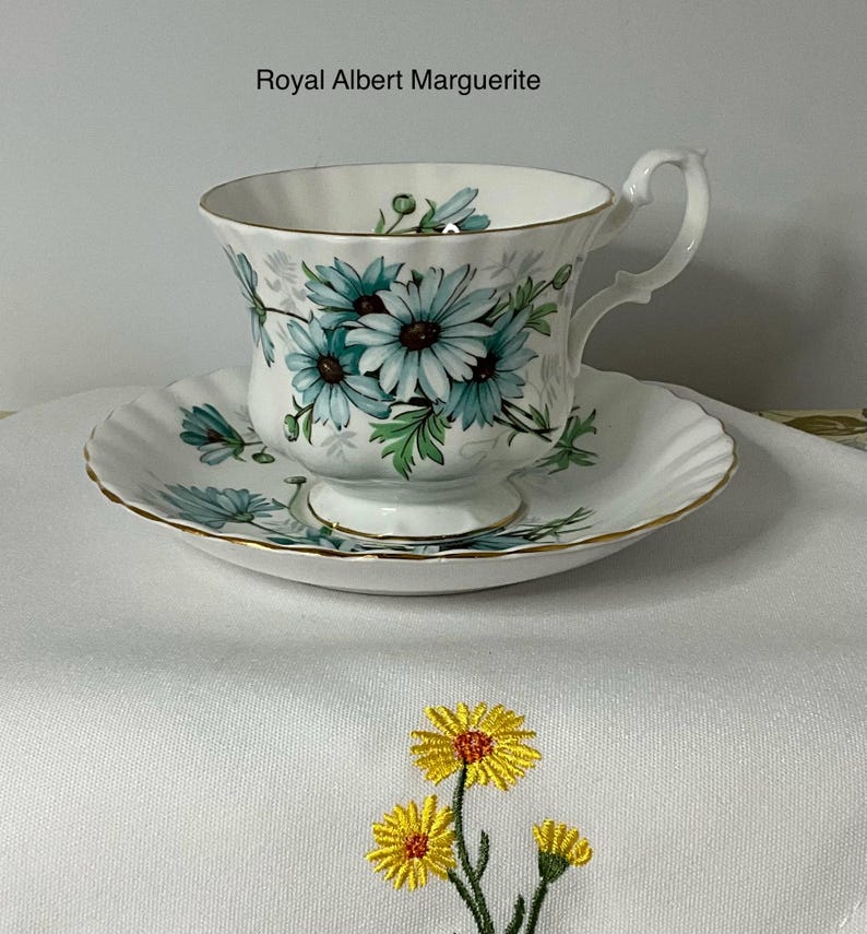 May include: A Royal Albert Marguerite teacup and saucer set featuring a white porcelain base with a blue daisy floral pattern and gold trim. The set is displayed above a white fabric cloth embroidered with three yellow daisy flowers.