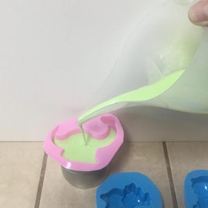 May include: Green liquid being poured into a pink dinosaur-shaped silicone mold. Two other molds, one blue and one pink, are visible in the background.