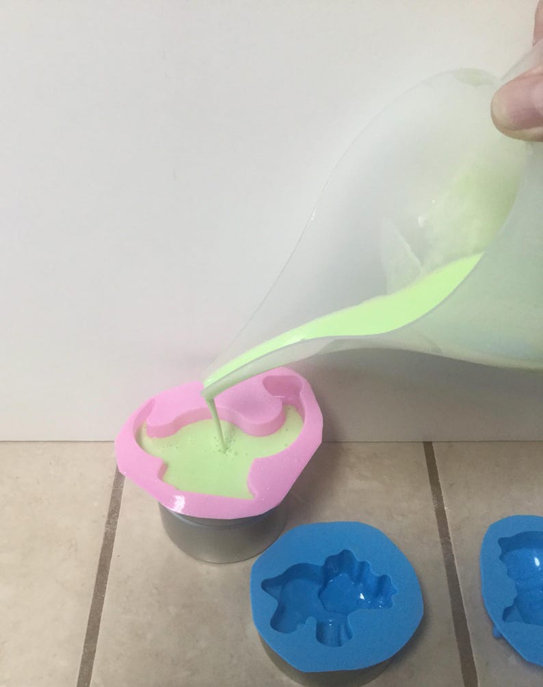 May include: Green liquid being poured into a pink dinosaur-shaped silicone mold. Two other molds, one blue and one pink, are visible in the background.