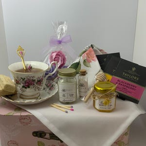 May include: A tea gift set featuring a floral porcelain teacup and saucer with a gold-tone spoon, a lavender soy candle, a small jar of honey with a bee charm, Taylors Blackberry & Raspberry tea bags, a rose-shaped soap, and a wooden honey dipper.