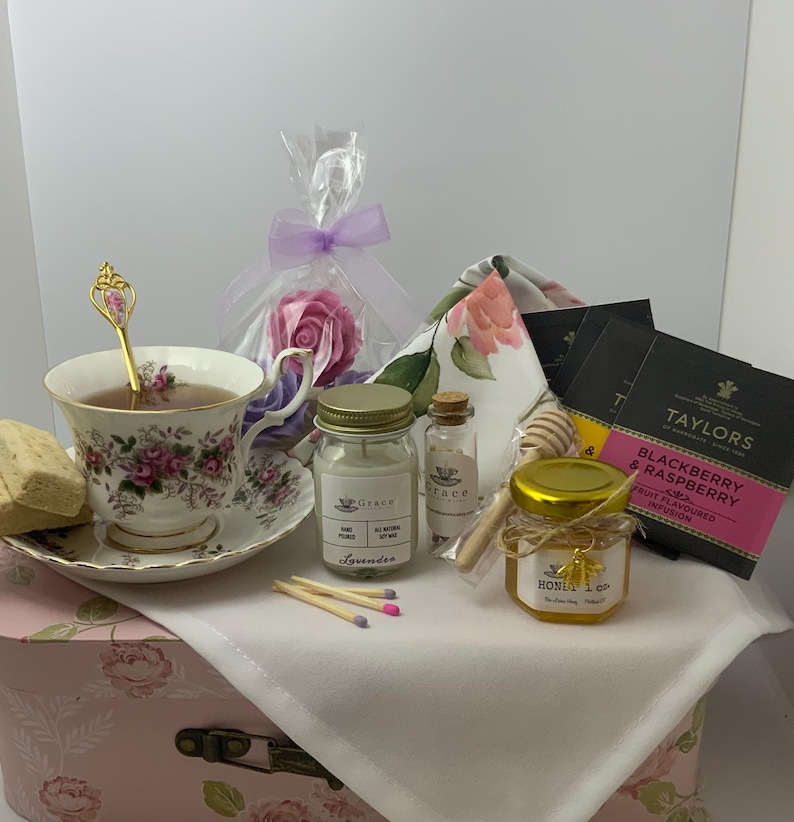 May include: A tea gift set featuring a floral porcelain teacup and saucer with a gold-tone spoon, a lavender soy candle, a small jar of honey with a bee charm, Taylors Blackberry & Raspberry tea bags, a rose-shaped soap, and a wooden honey dipper.
