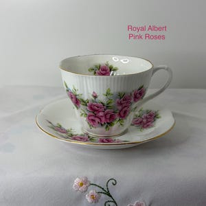 May include: A white porcelain teacup and saucer set featuring a ribbed texture, gold trim, and a floral pattern of pink roses and green leaves. The text Royal Albert Pink Roses is printed in pink on the upper right side of the image.