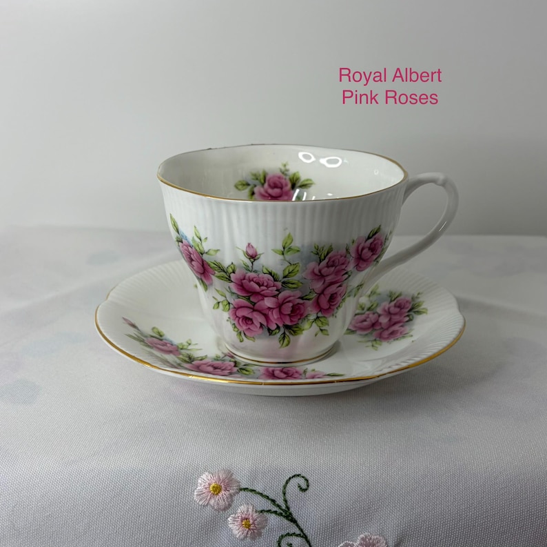 May include: A white porcelain teacup and saucer set featuring a ribbed texture, gold trim, and a floral pattern of pink roses and green leaves. The text Royal Albert Pink Roses is printed in pink on the upper right side of the image.