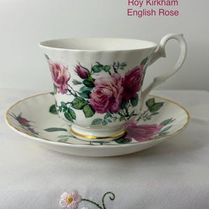 May include: A white porcelain teacup and saucer set featuring the Roy Kirkham English Rose pattern with pink roses, green leaves, and gold trim on the base and edges. The cup has a curved handle and a fluted rim, matching the saucer's design.