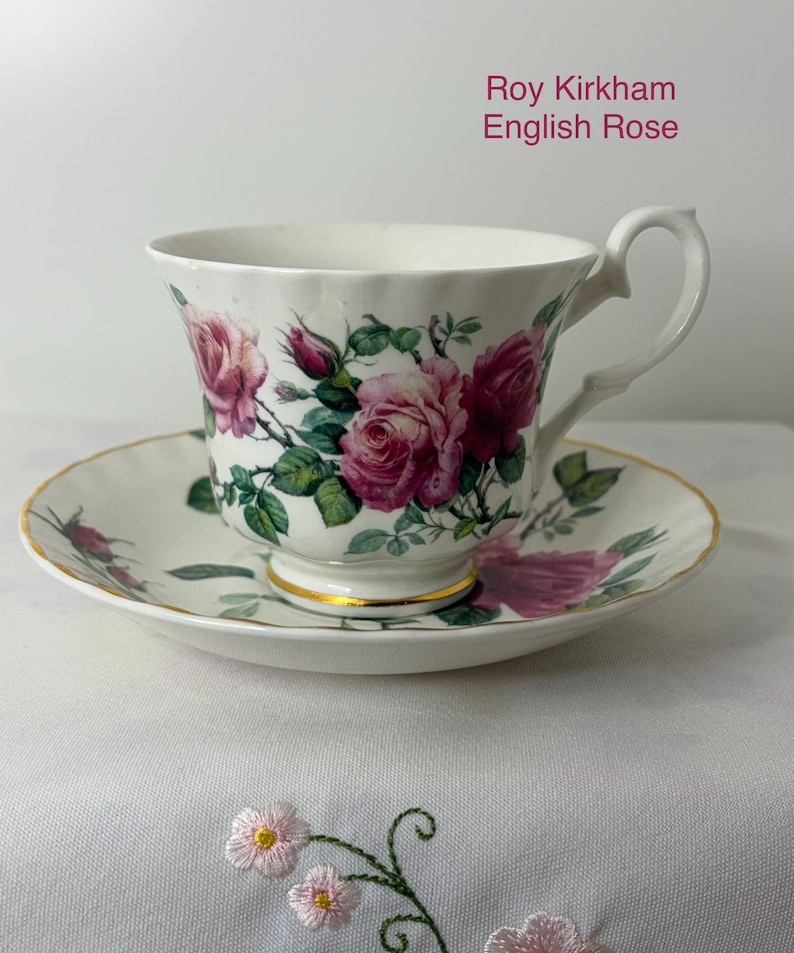 May include: A white porcelain teacup and saucer set featuring the Roy Kirkham English Rose pattern with pink roses, green leaves, and gold trim on the base and edges. The cup has a curved handle and a fluted rim, matching the saucer's design.